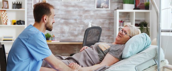 Male caregiver talking with a sick old lady lying in hospital bed, he takes the hand of the woman